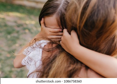 Sad dark-haired girl covers eyes with palm to wipe tears. Outdoor portrait of unhappy child with tanned skin crying and embracing mother's neck.