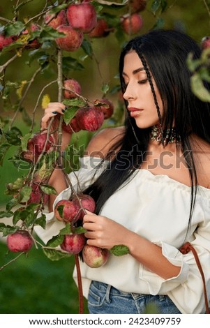 Sad, dark-haired woman in shoulderless white shirt, holding tree branches, laden with red apples. Close up of thoughtful pierced girl posing in apple orchard with closed eyes. Harvest, beauty concept.