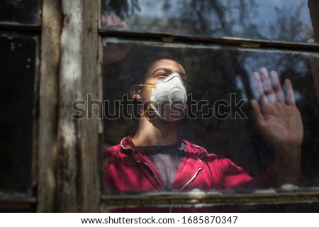 Sad depressed anxious young man wearing N95 protective medical mask looking through window,Coronavirus COVID-19 pandemic,self isolation quarantine,social physical distance,stay at home safe,hope faith