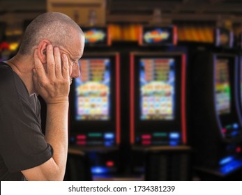 Sad And Depressed Caucasian Man Sitting With His Hands On His Head In Front Of Rows Of Casino Slot Machines. Gambling Addiction Theme Image.  Close Up Portrait.