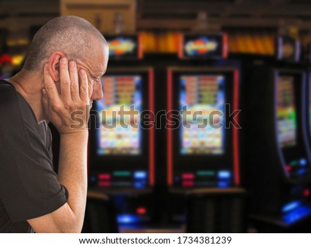 Sad and depressed caucasian man sitting with his hands on his head in front of rows of casino slot machines. Gambling addiction theme image.  Close up portrait.