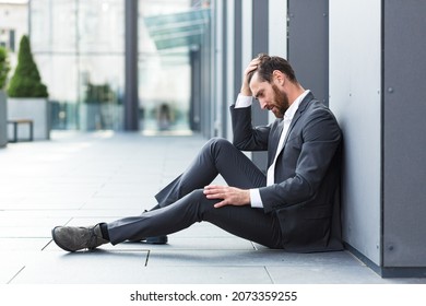 Sad depressed entrepreneur in formal suit worker man sitting near outdoors street near modern office business center. Upset male businessman lost job due financial crisis employee has problem. outside