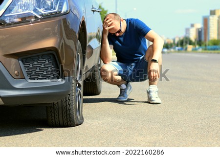 A sad and depressed man is sitting next to a brown car with a punctured tire.