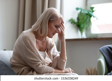 Sad Depressed Middle Aged Mature Woman Sitting Alone At Home Touching Head Feeling Headache Migraine, Upset Tired Old Senior Lady Widow Mourning Suffer From Grief Loneliness Mental Problems Concept