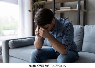 Sad depressed millennial man sitting on sofa in living room with head-down, suffers due to personal life troubles, goes through break up, split, divorce, after quarrel mood, feeling remorse concept