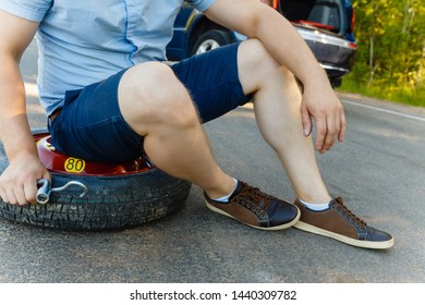 Sad And Depressed Person Sits On A Spare Wheel Near A Blue Car With A Punctured Tire And An Open Trunk. A Man Calls Using A Smartphone Mobile Tire Service.