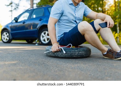 Sad And Depressed Person Sits On A Spare Wheel Near A Blue Car With A Punctured Tire And An Open Trunk. A Man Calls Using A Smartphone Mobile Tire Service.