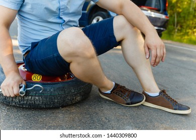 Sad And Depressed Person Sits On A Spare Wheel Near A Blue Car With A Punctured Tire And An Open Trunk. A Man Calls Using A Smartphone Mobile Tire Service.