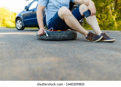 Sad And Depressed Person Sits On A Spare Wheel Near A Blue Car With A Punctured Tire And An Open Trunk. A Man Calls Using A Smartphone Mobile Tire Service.