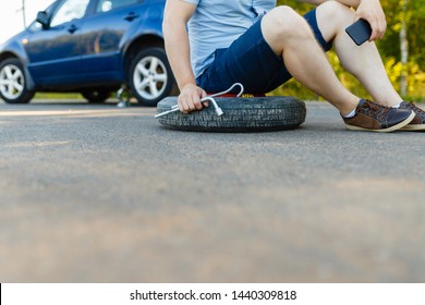 Sad And Depressed Person Sits On A Spare Wheel Near A Blue Car With A Punctured Tire And An Open Trunk. A Man Calls Using A Smartphone Mobile Tire Service.