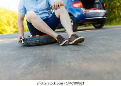 Sad And Depressed Person Sits On A Spare Wheel Near A Blue Car With A Punctured Tire And An Open Trunk. A Man Calls Using A Smartphone Mobile Tire Service.