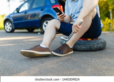 Sad And Depressed Person Sits On A Spare Wheel Near A Blue Car With A Punctured Tire And An Open Trunk. A Man Calls Using A Smartphone Mobile Tire Service.