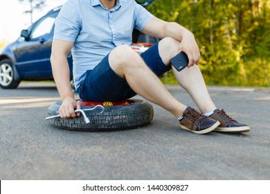 Sad And Depressed Person Sits On A Spare Wheel Near A Blue Car With A Punctured Tire And An Open Trunk. A Man Calls Using A Smartphone Mobile Tire Service.