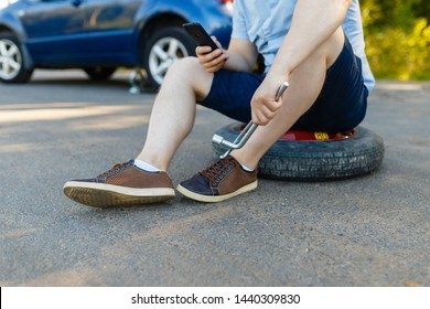 Sad And Depressed Person Sits On A Spare Wheel Near A Blue Car With A Punctured Tire And An Open Trunk. A Man Calls Using A Smartphone Mobile Tire Service.