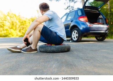 Sad and depressed person sits on a spare wheel near a blue car with a punctured tire and an open trunk. A man calls using a smartphone mobile tire service. - Shutterstock ID 1440309833