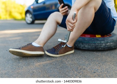 Sad And Depressed Person Sits On A Spare Wheel Near A Blue Car With A Punctured Tire And An Open Trunk. A Man Calls Using A Smartphone Mobile Tire Service.