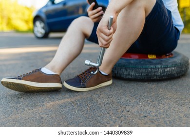 Sad And Depressed Person Sits On A Spare Wheel Near A Blue Car With A Punctured Tire And An Open Trunk. A Man Calls Using A Smartphone Mobile Tire Service.