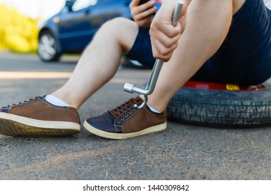 Sad And Depressed Person Sits On A Spare Wheel Near A Blue Car With A Punctured Tire And An Open Trunk. A Man Calls Using A Smartphone Mobile Tire Service.