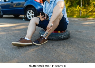 Sad And Depressed Person Sits On A Spare Wheel Near A Blue Car With A Punctured Tire And An Open Trunk. A Man Calls Using A Smartphone Mobile Tire Service.