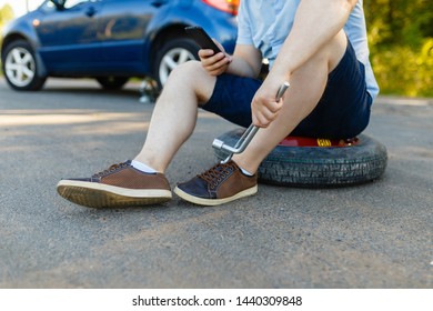 Sad And Depressed Person Sits On A Spare Wheel Near A Blue Car With A Punctured Tire And An Open Trunk. A Man Calls Using A Smartphone Mobile Tire Service.