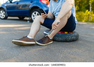 Sad And Depressed Person Sits On A Spare Wheel Near A Blue Car With A Punctured Tire And An Open Trunk. A Man Calls Using A Smartphone Mobile Tire Service.