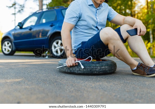 Sad and depressed person sits on a
spare wheel near a blue car with a punctured tire and an open
trunk. A man calls using a smartphone mobile tire
service.