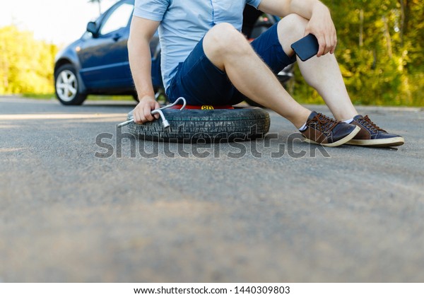 Sad and depressed person sits on a\
spare wheel near a blue car with a punctured tire and an open\
trunk. A man calls using a smartphone mobile tire\
service.