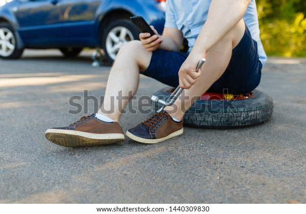 Sad and depressed person sits on a\
spare wheel near a blue car with a punctured tire and an open\
trunk. A man calls using a smartphone mobile tire\
service.
