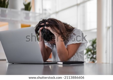 sad depressed remote working woman sitting disappointed with headache infront of a laptop or notebook on her work desk in modern airy bright living room home office with many windows
