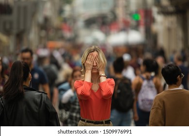 Sad depressed woman covers his eyes with his hands surrounded by people walking in crowded street. Panic attack in public place. 