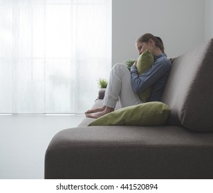 Sad Depressed Woman At Home, She Is Sitting On The Couch And Hugging A Pillow, Loneliness And Sadness Concept