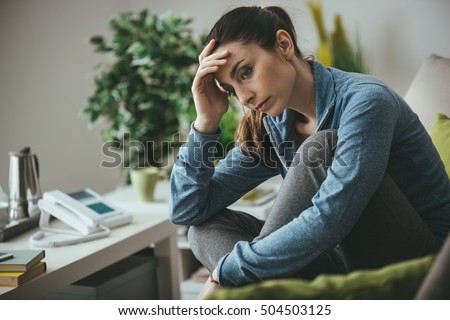 Sad depressed woman at home sitting on the couch, looking down and touching her forehead, loneliness and pain concept