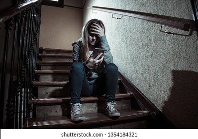 Sad Depressed Young Teenager Girl Victim Of Cyberbullying By Mobile Smart Phone Siting On Stairs Feeling Lonely, Unhappy, Hopeless And Abused. Bullied By Text Message On Social Media App. Dark Light