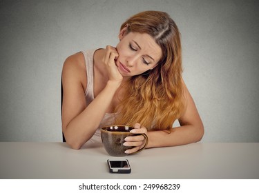 Sad depressed young woman sitting at table drinking coffee looking at her mobile phone waiting for a call text message isolated on grey wall background.