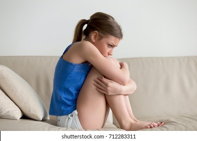 Sad Depressed Young Woman Sitting On Sofa Embracing Her Knees, Upset Heartbroken Teen Girl Feeling Vulnerable Ashamed, Frustrated Adolescent Teenager Having Problem, Suffering From Bullying Or Abuse