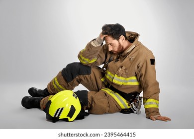 Sad, despaired firefighter crying, rubbing forehead, while sitting on floor. Side view of pensive, exhausted fireman resting, after fighting fire, on gray studio background. Concept of emotions, work. - Shutterstock ID 2395464415