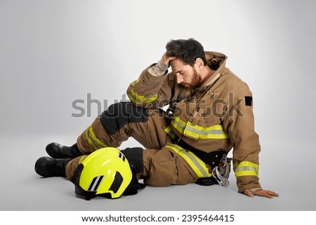 Sad, despaired firefighter crying, rubbing forehead, while sitting on floor. Side view of pensive, exhausted fireman resting, after fighting fire, on gray studio background. Concept of emotions, work.