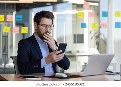 Sad disappointed businessman reading news on phone, man in business suit using app on smartphone, looking upset, received bad news online in email, working with laptop inside office. - Powered by Shutterstock