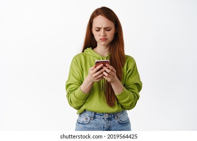 Sad And Disappointed Ginger Teen Girl Looking Upset At Her Phone, Staring At Smartphone With Regret Or Jealousy, Standing Against White Background