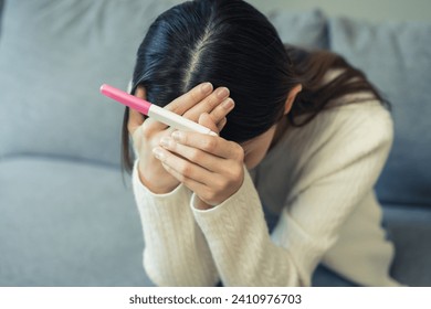 Sad, Disappointed motherhood asian young woman hand holding pregnancy test, upset ovulation problem, anxiety after result at positive sitting on couch at home. Unwanted Pregnant, contraception concept - Powered by Shutterstock