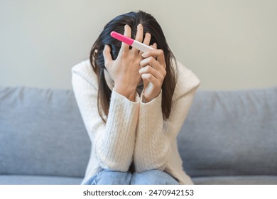 Sad, Disappointed motherhood asian young woman hand holding pregnancy test, upset ovulation problem, anxiety after result at positive sitting on couch at home. Unwanted Pregnant, contraception concept - Powered by Shutterstock