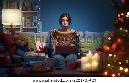 Sad disappointed woman opening Christmas gifts at home, she has received an ugly Christmas sweater