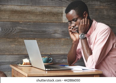 Sad And Disappointed Young Black Freelancer In Glasses And Pink Shirt Sitting At The Table At A Cafe, Checking Voicemail On Mobile Phone And Finding Out That The Investor Cancelled The Meeting