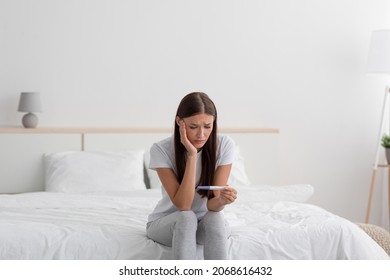 Sad Disappointment Young European Woman Hold Pregnancy Test, Sits On Bed Alone In Bedroom Interior. Infertility, Female Health Problems, Unwanted Pregnancies And Loneliness After Breakup, Free Space