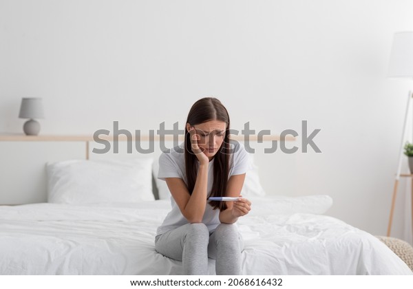 Sad disappointment young european woman hold\
pregnancy test, sits on bed alone in bedroom interior. Infertility,\
female health problems, unwanted pregnancies and loneliness after\
breakup, free space