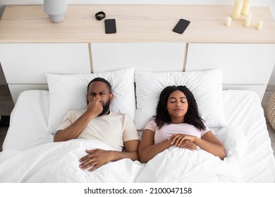 Sad Disgruntled Millennial African American Husband Closes Nose From Bad Scent, Wife Sleeps On White Bed Together In Bedroom Interior, Top View. Smell And Problems At Home, Sleeplessness, Free Space