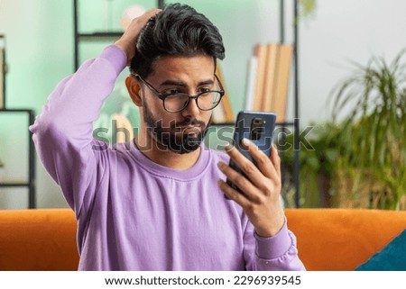 Sad displeased indian man use smartphone typing browsing, loses becoming surprised sudden lottery results, bad news, fortune loss, fail, deadline, virus. Young hindu guy at home living room on couch