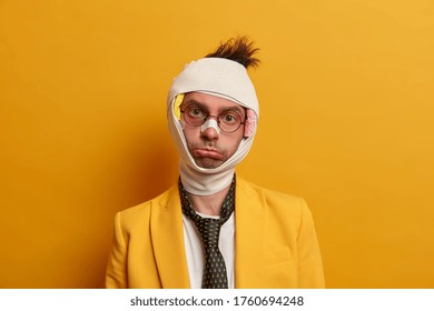 Sad displeased man with gloomy expression looks distressed at camera, has bad mood after accident happened with him, wears plaster on broken nose, big hematoma under eye, isolated on yellow wall