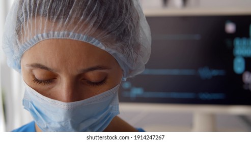 Sad Doctor In Safety Mask And Hat Standing Near Monitor With No Heart Rate. Close Up Of Upset Woman Medical Worker Stressed About Death Of Patient With No Heart Rate On Ecg Monitor On Background