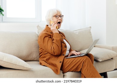 A Sad, Elderly Businesswoman Is Sitting On A Sofa In A Cozy Apartment In A Brown Suit Working From Home And Talking On The Phone Loudly Shouts With Her Mouth Wide Open